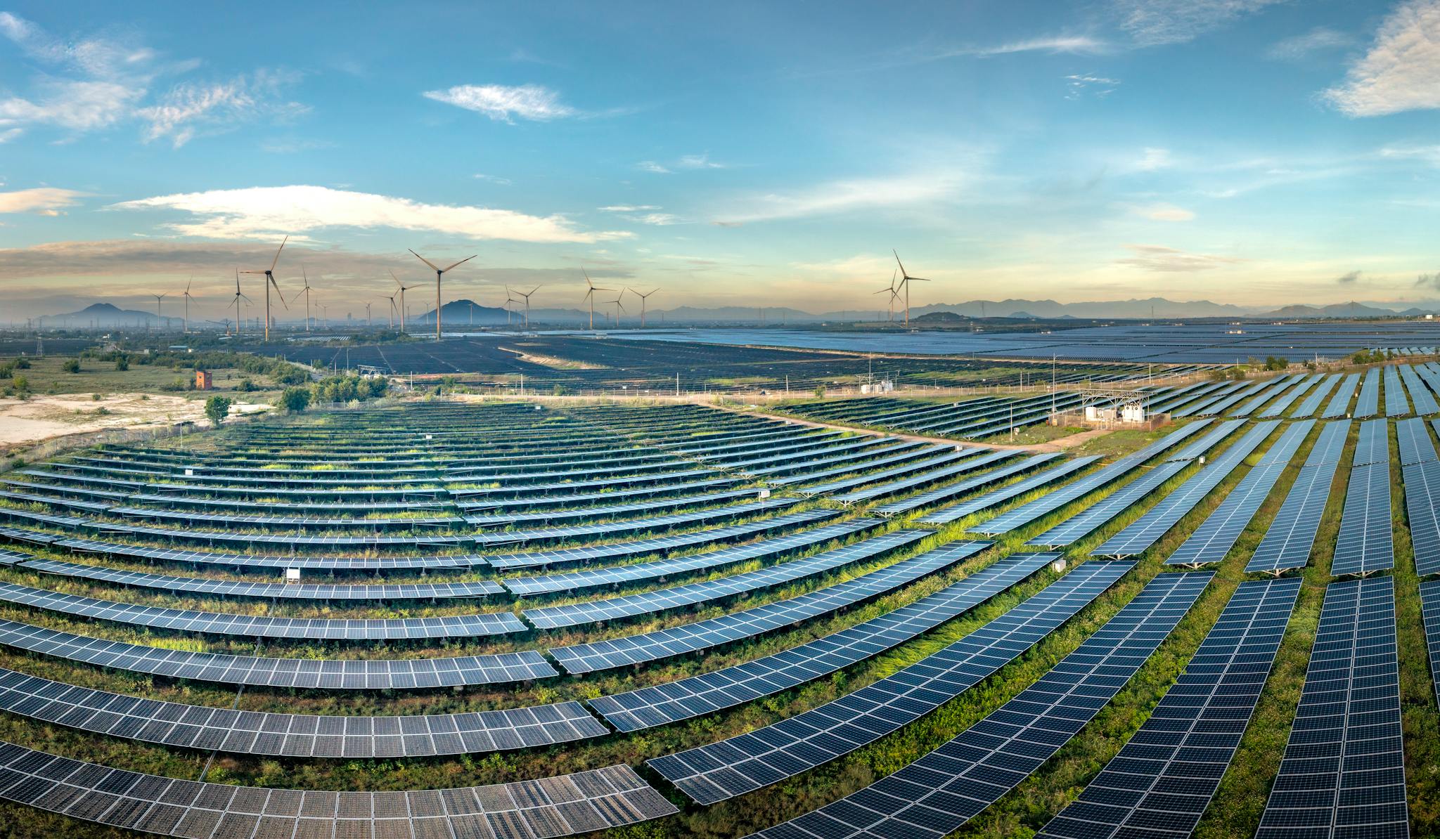 Expansive solar farm with wind turbines in the background under a clear blue sky.
